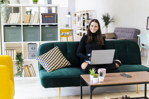 Young female blogger sitting with laptop on sofa at loft apartment - GIOF09425