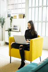 Smiling beautiful young woman using laptop for blogging while sitting on chair at home - GIOF09429