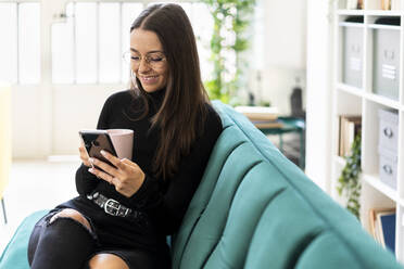 Smiling beautiful female influencer using phone while sitting with coffee cup on sofa at home - GIOF09432