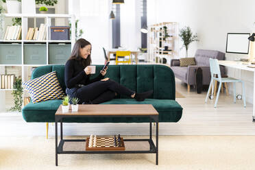 Smiling female blogger using phone while sitting with coffee cup on sofa at home - GIOF09433