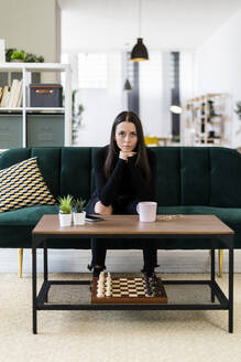 Confident young woman sitting on sofa in living room at loft apartment - GIOF09435