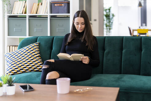 Beautiful young woman reading book while sitting on sofa at home - GIOF09437