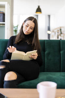 Beautiful young woman with long brown hair reading book while sitting on sofa at home - GIOF09438