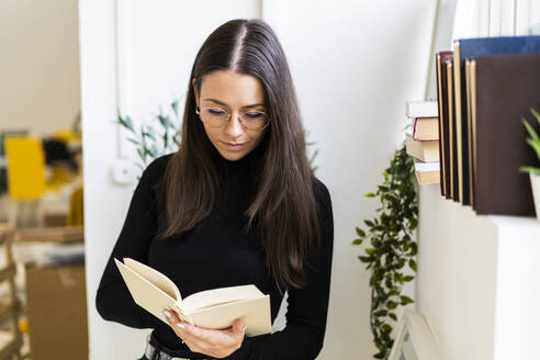 Beautiful female brunette reading book while standing at home - GIOF09441