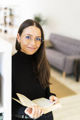 Lächelnde schöne junge Frau, die ein Buch hält, während sie zu Hause am Regal steht - GIOF09443