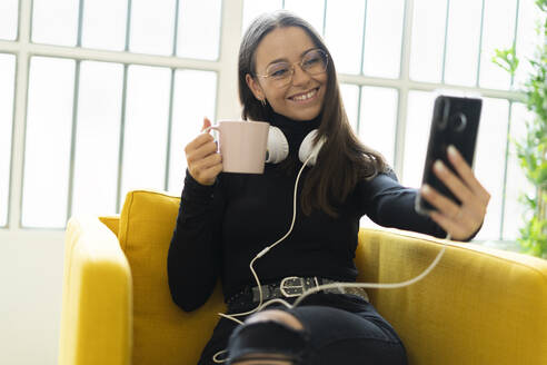 Smiling young female blogger taking selfie with coffee cup and headphones against window at home - GIOF09444