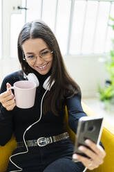 Smiling female blogger taking selfie with coffee cup and headphones at home - GIOF09445