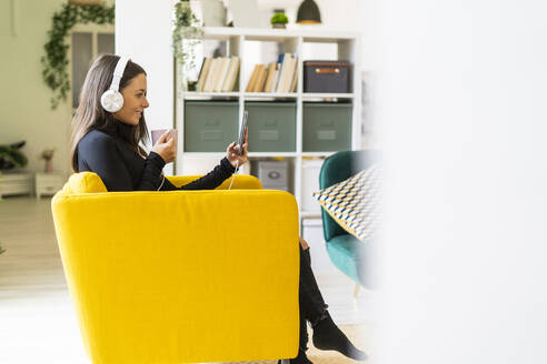 Smiling female blogger taking selfie with coffee cup and headphones while sitting on yellow chair at loft apartment - GIOF09446