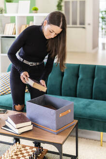 Beautiful young female brunette collecting books in box on coffee table at loft apartment - GIOF09447