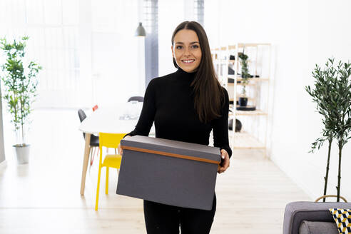 Smiling beautiful young woman carrying box at loft apartment - GIOF09452