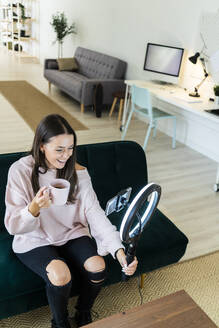 Smiling young female influencer sitting on sofa vlogging through mobile phone while holding coffee cup at loft apartment - GIOF09461