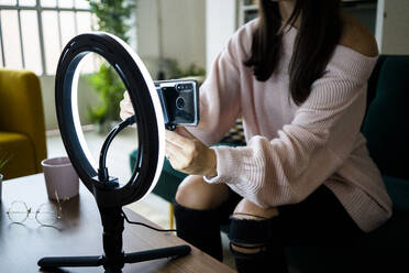 Young female influencer sitting on sofa while adjusting smart phone on lighting equipment for vlogging at loft apartment - GIOF09465