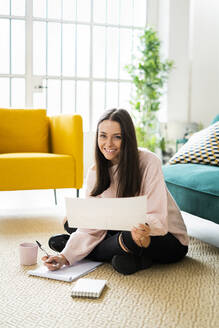 Happy beautiful young woman sitting with note pads and coffee cup on carpet against sofa at home - GIOF09472
