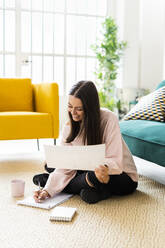 Lächelnde junge Frau, die in einen Notizblock schreibt, während sie mit einer Kaffeetasse auf dem Teppich vor dem Sofa in einer Loftwohnung sitzt - GIOF09473