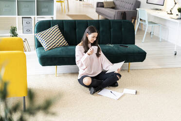 Schöne junge Frau trinkt Kaffee beim Lesen von Papier durch Notizblöcke auf Teppich gegen Sofa zu Hause - GIOF09474