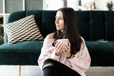Nachdenkliche junge Frau, die wegschaut, während sie mit einer Kaffeetasse auf dem Sofa zu Hause sitzt - GIOF09475