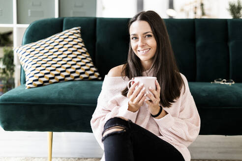 Smiling woman sitting with coffee cup against sofa at home - GIOF09476