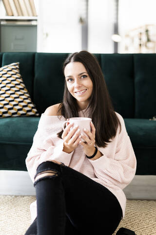 Happy young woman sitting with coffee cup against sofa at home stock photo