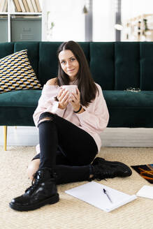 Beautiful young woman sitting with coffee cup and note pads on carpet against sofa at home - GIOF09478
