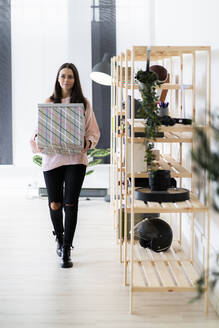 Beautiful young woman carrying box while walking by rack at loft apartment - GIOF09479