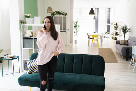 Happy beautiful young woman talking on mobile phone while standing in living room at loft apartment stock photo