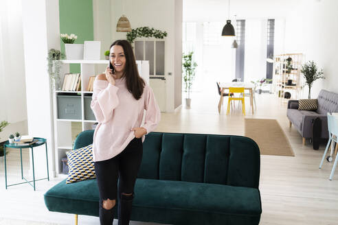 Happy beautiful young woman talking on mobile phone while standing in living room at loft apartment - GIOF09482