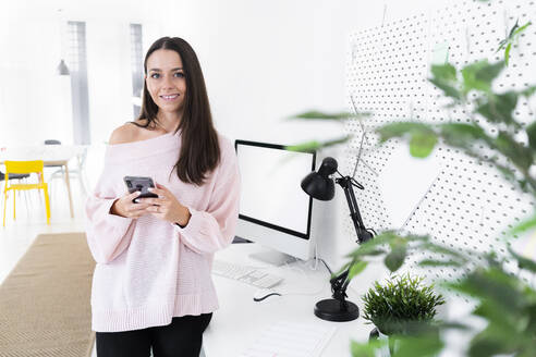 Smiling young female influencer standing with mobile phone at computer desk in loft apartment - GIOF09488