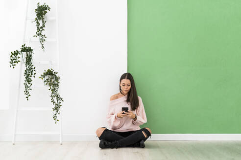 Beautiful young woman text messaging through mobile phone while sitting on floor by ladder against wall - GIOF09492