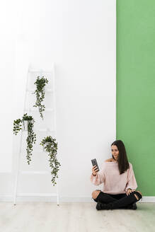 Young woman taking selfie through smart phone while sitting on floor by ladder against wall - GIOF09493