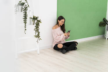Lächelnde Frau, die eine Textnachricht über ihr Mobiltelefon sendet, während sie auf dem Boden neben einer Leiter an der Wand sitzt - GIOF09494
