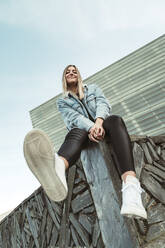 Lächelnde junge Frau sitzt am Rande einer Stützmauer gegen den Himmel - MTBF00687