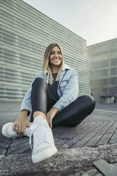 Smiling beautiful woman looking away while sitting on footpath - MTBF00688