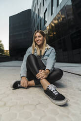 Smiling young woman looking away while sitting on footpath against modern building - MTBF00694