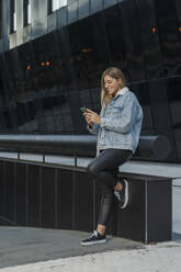 Lächelnde junge Frau, die ihr Smartphone benutzt, während sie sich an ein Geländer vor einem modernen Gebäude lehnt - MTBF00698