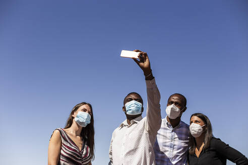 Business professionals taking selfie with protective face masks against clear blue sky on sunny day - LJF01792