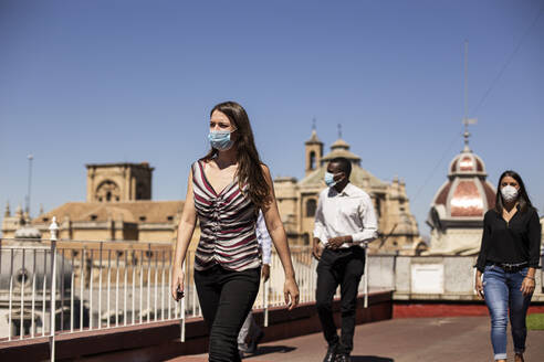 Business people wearing masks while walking at rooftop of office building - LJF01793