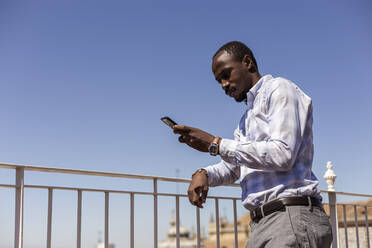 Männlicher Unternehmer, der sein Smartphone benutzt, während er sich gegen den klaren blauen Himmel auf ein Geländer stützt - LJF01797