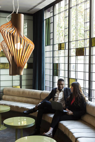 Male and female colleagues discussing over laptop while sitting on sofa in modern bar stock photo