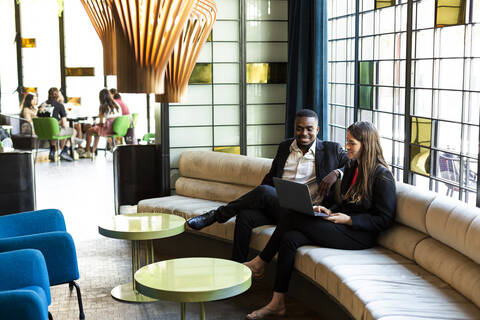 Smiling male and female business professionals discussing over laptop while sitting on sofa in modern bar stock photo