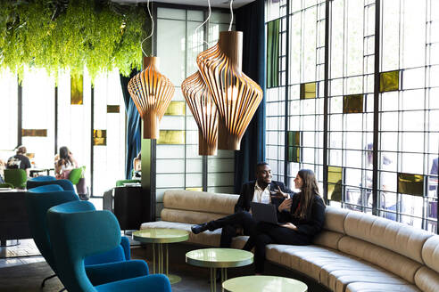 Multi-ethnic male and female colleagues discussing while sitting on sofa in modern bar - LJF01807