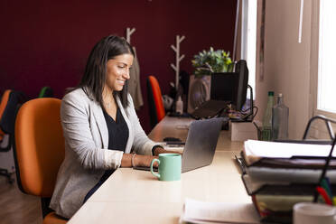 Smiling businesswoman using laptop at desk in coworking office - LJF01818