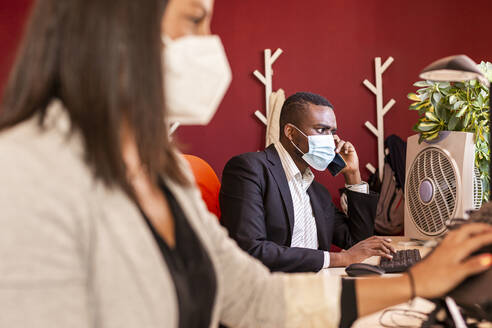 Young businessman in protective face mask talking on mobile phone at office during COVID-19 - LJF01830