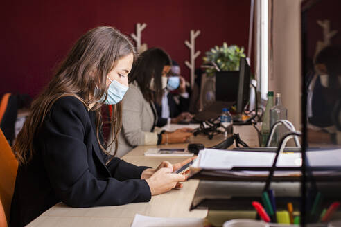 Young businesswoman in protective face mask text messaging through smart phone at desk in office during pandemic - LJF01832