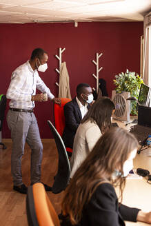 Male and female colleagues in protective face masks working at desk in coworking office during COVID-19 - LJF01833