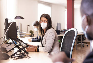 Female business in face mask looking at colleague while sitting at desk in office during COVID-19 - LJF01838