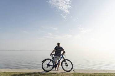 Man standing with bicycle looking at sea against blue sky - CHPF00691