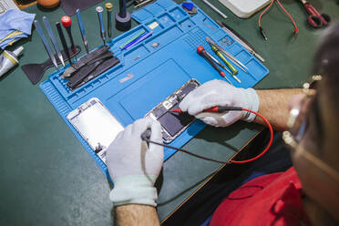 Male engineer examining damaged smart phone at workbench at repair shop - LJF01841