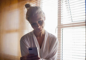 Blonde Frau im Morgenmantel, die zu Hause eine Textnachricht über ihr Mobiltelefon sendet - AJOF00468