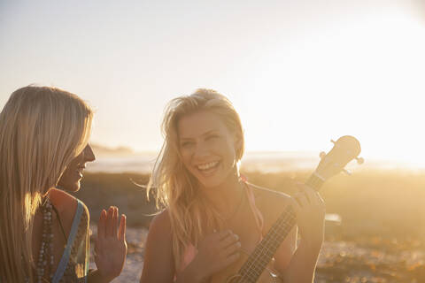 Cheerful young woman playing ukulele while female friend singing at beach during sunset stock photo