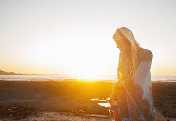Junge Frau hört Musik über Kopfhörer, während sie am Strand gegen den Himmel bei Sonnenuntergang steht - AJOF00480